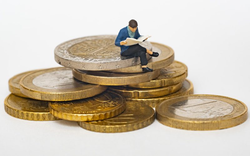 Miniature person sitting on stack of coins reading newspaper